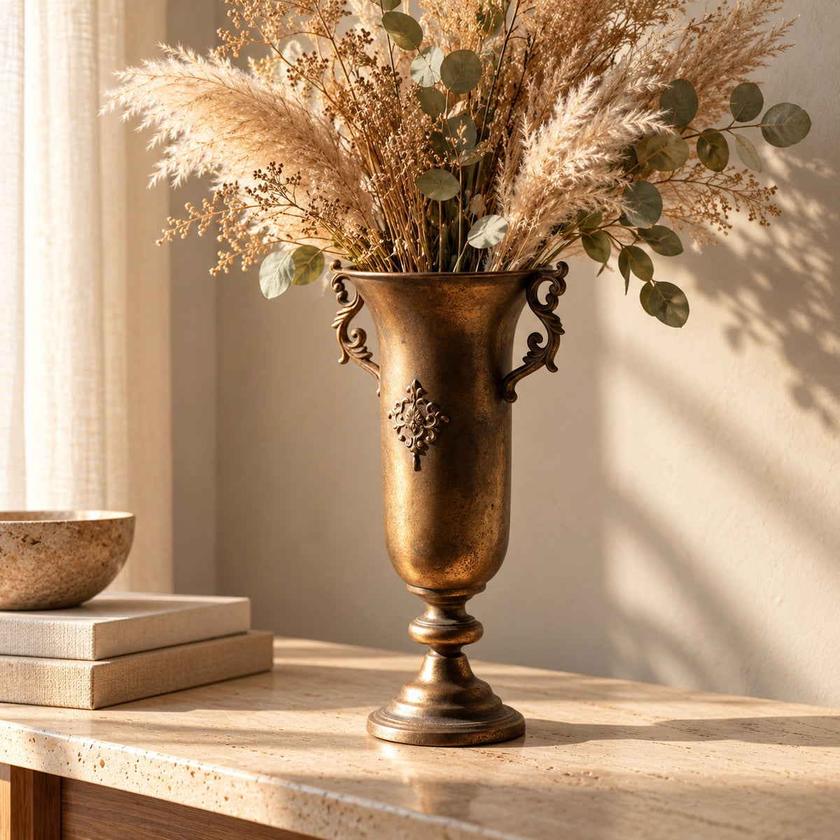 Antique gold trumpet vase with scroll handles holding pampas grass on a travertine console