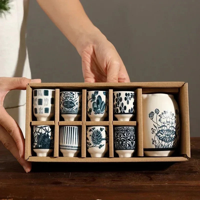 A blue and white ceramic sake set in a kraft box: eight small cups in different patterns and a tall lotus-pattern decanter.