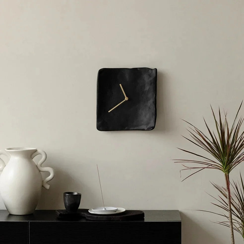 Black crumpled resin wall clock with gold-toned hands, hung above a black sideboard with a white vase and a plant