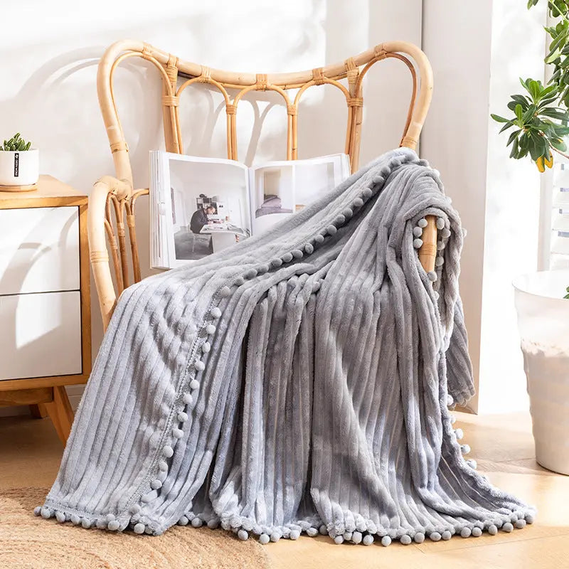 A gray flannel throw with raised stripes and a pom-pom edge in the same gray, draped over a rattan armchair