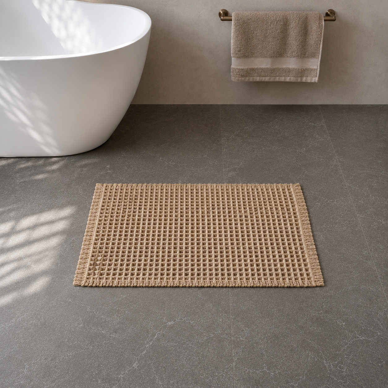 Khaki waffle weave bath mat with fringed edges on a dark grey stone floor beside a freestanding bath