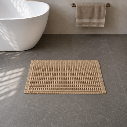 Khaki waffle weave bath mat with fringed edges on a dark grey stone floor beside a freestanding bath