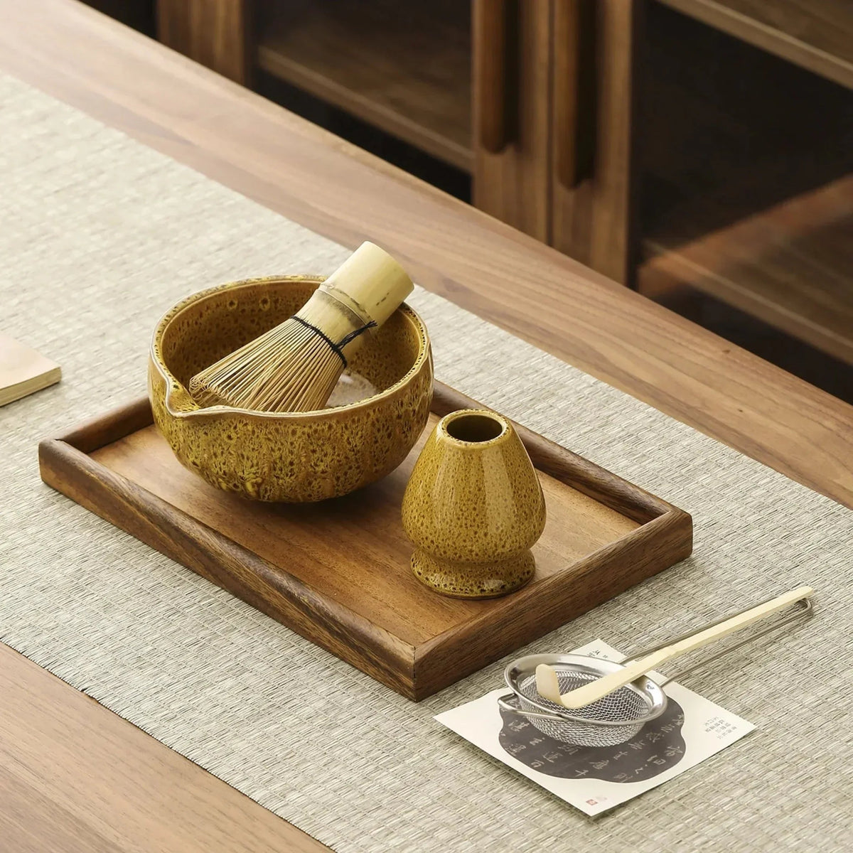 Yellow speckled ceramic matcha bowl with a bamboo whisk resting in it, matching whisk stand, steel sifter and bamboo scoop, shown on a wooden tray that is not included