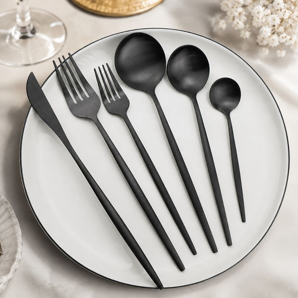 Matte black cutlery place setting on a white plate: dinner knife, dinner fork, dessert fork, dinner spoon, dessert spoon and teaspoon