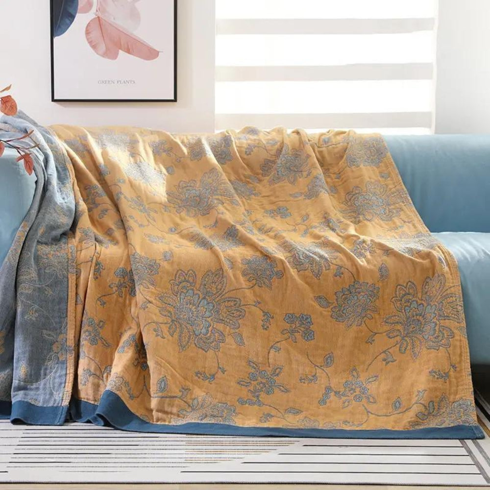 Mustard floral jacquard throw blanket draped over a pale aqua sofa, blue flowers woven across a mustard face with a deep blue border