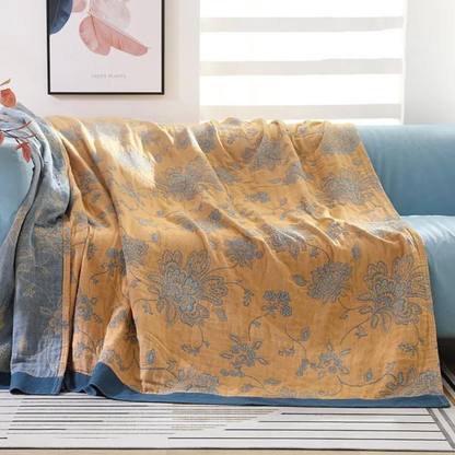 Mustard floral jacquard throw blanket draped over a pale aqua sofa, blue flowers woven across a mustard face with a deep blue border