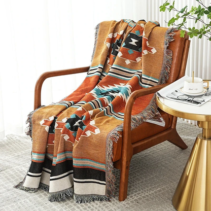 A mustard and rust jacquard throw with black and teal stepped medallions, draped over a wooden armchair beside a gold side table