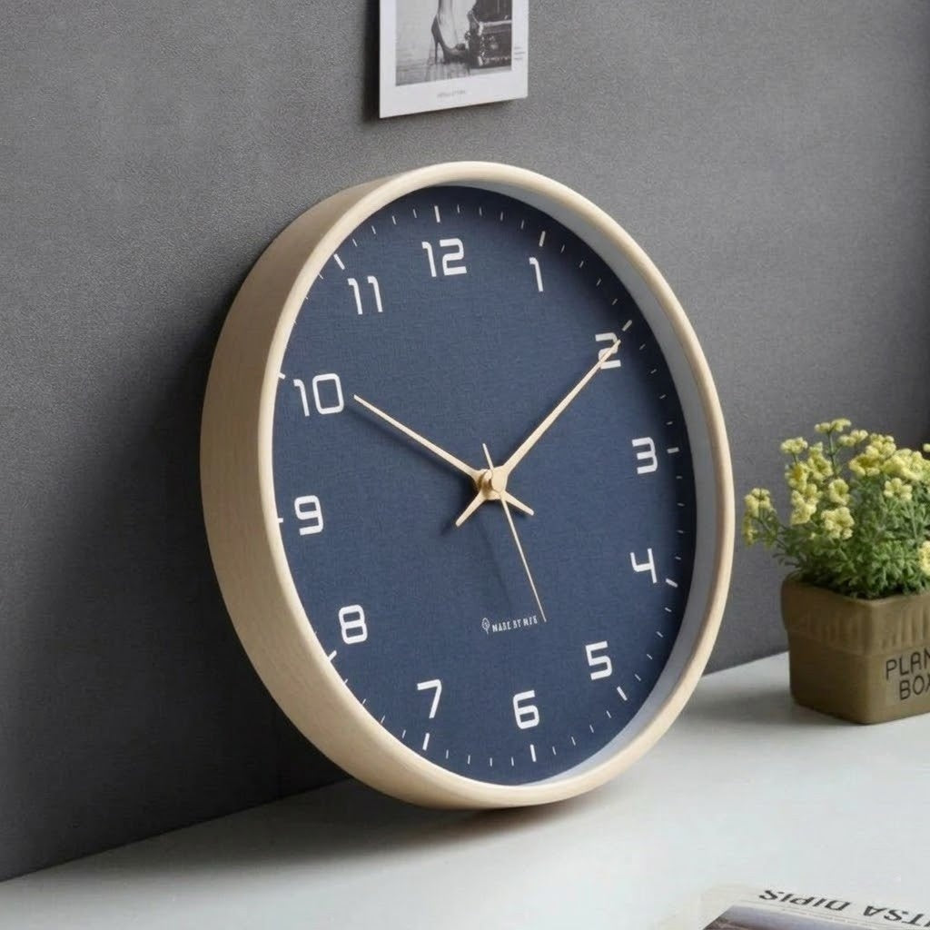 Navy blue wall clock with white numbers and a pale wood frame resting against a dark gray wall on a white desk