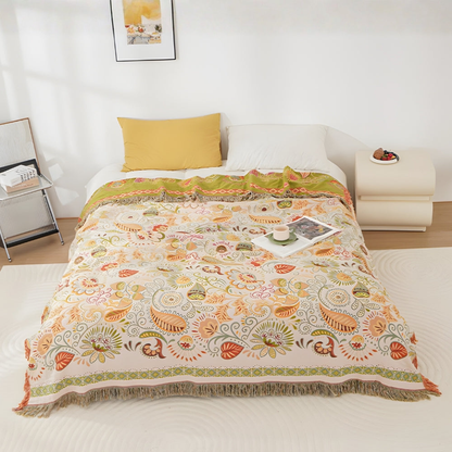 An orange and cream leaf and paisley throw blanket spread over a white bed, with the top edge turned back to show its olive reverse and tassels along the foot