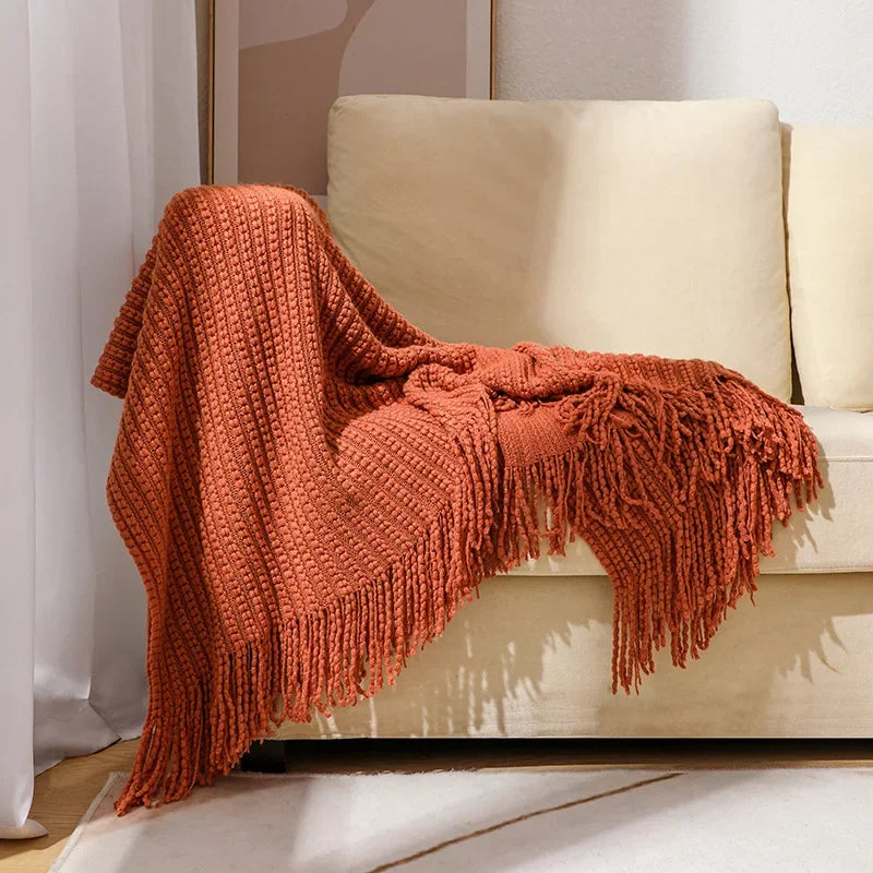 A rust bobble knit throw blanket with fringed ends draped over the arm of a cream sofa.