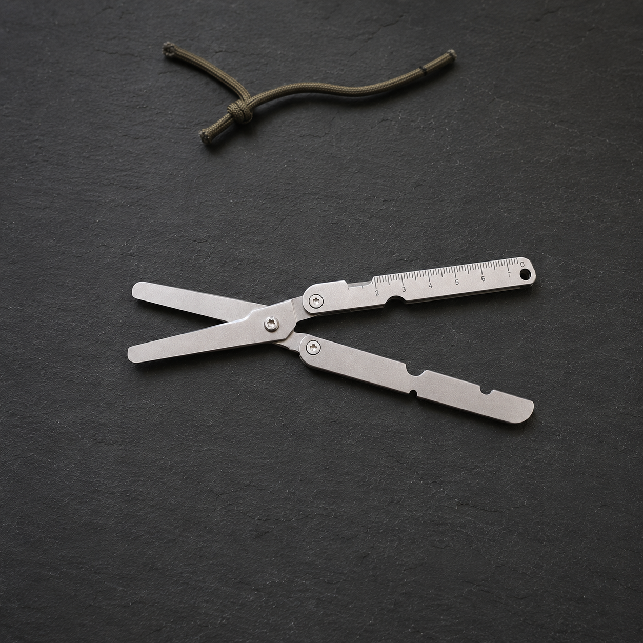Silver folding pocket scissors lying open on a dark slate worktop, a short length of cord beside them.