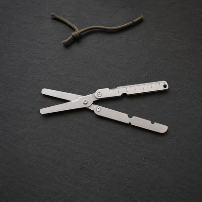 Silver folding pocket scissors lying open on a dark slate worktop, a short length of cord beside them.