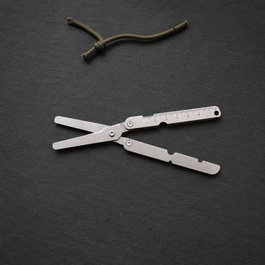 Silver folding pocket scissors lying open on a dark slate worktop, a short length of cord beside them.