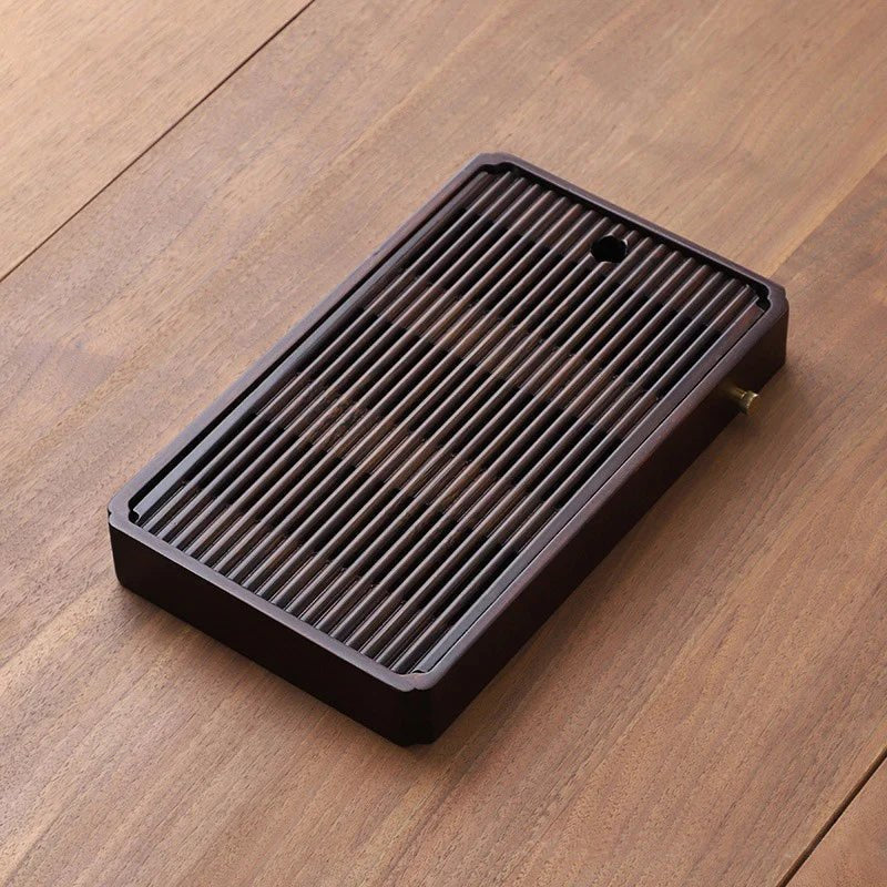 Small slatted bamboo tea tray in a dark brown finish on a wooden table, a gold-toned drain cap on its long side.