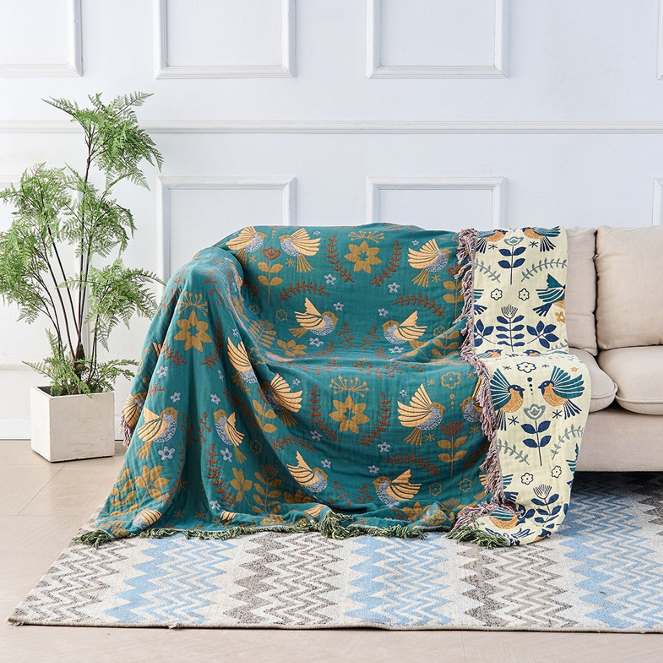 A teal cotton throw woven with golden birds draped over a cream sofa, with its cream reverse and navy birds showing down one side
