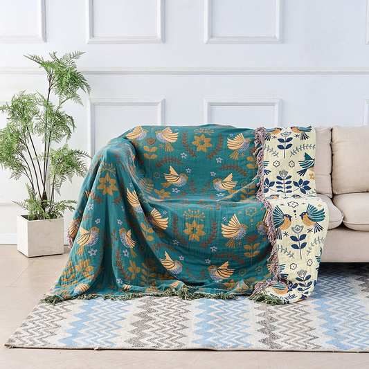 A teal cotton throw woven with golden birds draped over a cream sofa, with its cream reverse and navy birds showing down one side