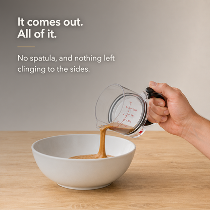 A hand tilting the clear measuring cup mouth-down over a white mixing bowl on an oak worktop, one continuous column of spread leaving the mouth as the plate inside pushes it out, the inside of the cup left bare