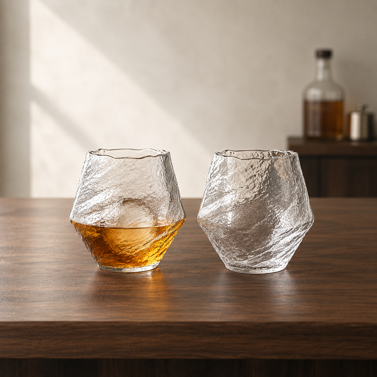 Two hammered whiskey tumblers on a walnut bar top, one filled with whiskey over ice