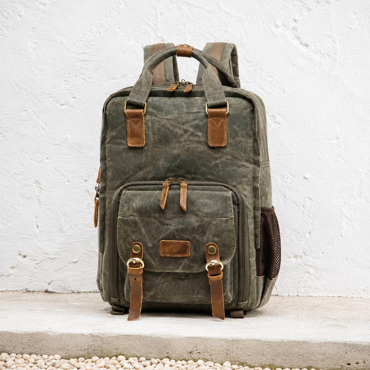Army green waxed canvas camera backpack against a whitewashed courtyard wall.