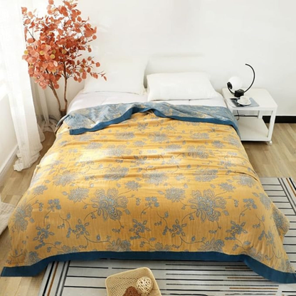 Mustard cotton gauze throw blanket spread flat across a bed in a bright bedroom, the floral pattern covering the whole top