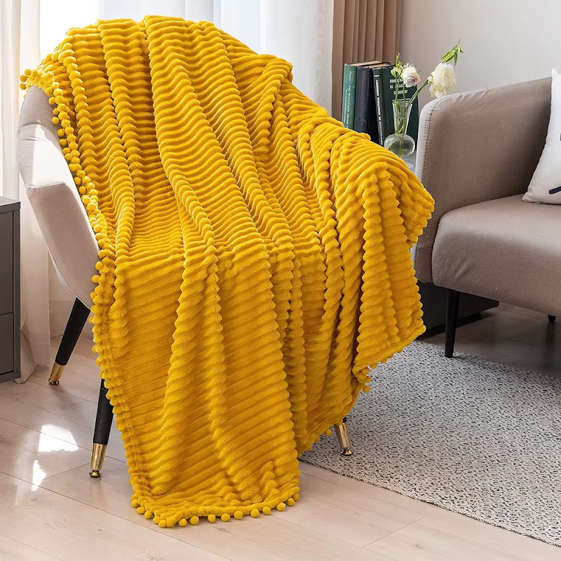 A mustard flannel throw with raised stripes and matching pom-poms hanging over a white armchair