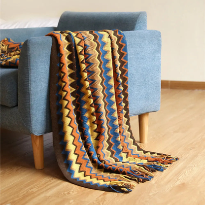 An orange and blue chevron throw draped over the arm of a blue couch, its tassels resting on a wooden floor.