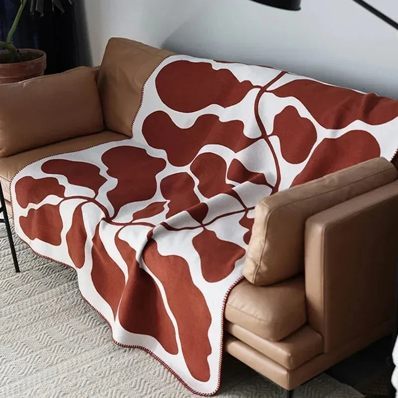 A rust and off-white abstract pattern throw blanket spread across the seat and arm of a tan leather sofa.