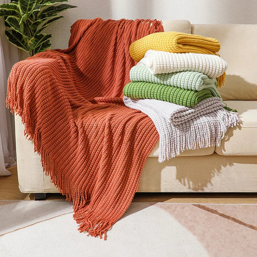 A rust bobble knit throw blanket thrown over a sofa, with mustard, cream, sage, green and gray throws folded on the seat.