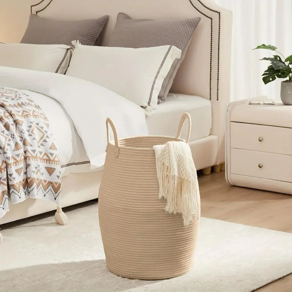 A tan rope laundry hamper with a cream throw hanging over the rim, beside a bed.