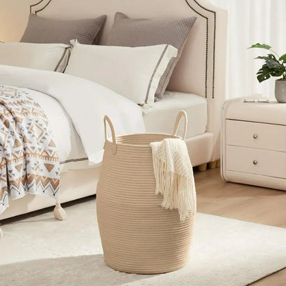 A tan rope laundry hamper with a cream throw hanging over the rim, beside a bed.