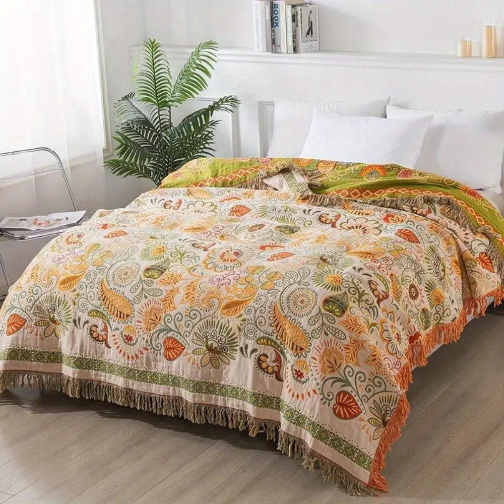 A cream throw blanket patterned with orange and gold leaves and paisleys over a bed in a bright bedroom, its olive underside folded back at the head and tassels hanging at the foot and side