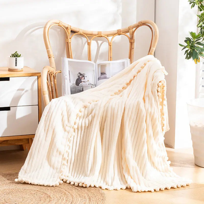 A cream flannel throw with raised stripes and matching pom-poms over a rattan armchair beside a white drawer unit