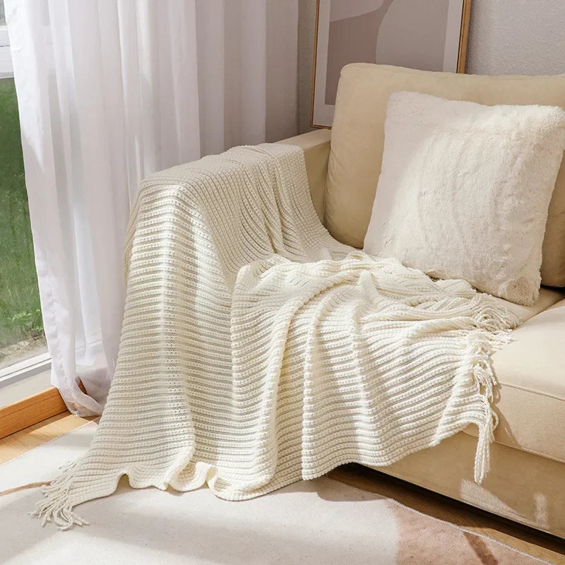 A cream bobble knit throw blanket with fringed ends spread over a sofa beside a sunny window.