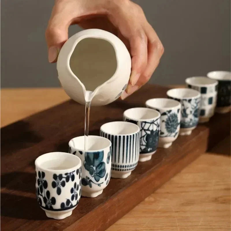 The decanter pouring a thin stream from its lip into the first of eight blue and white cups lined up along a wooden board.