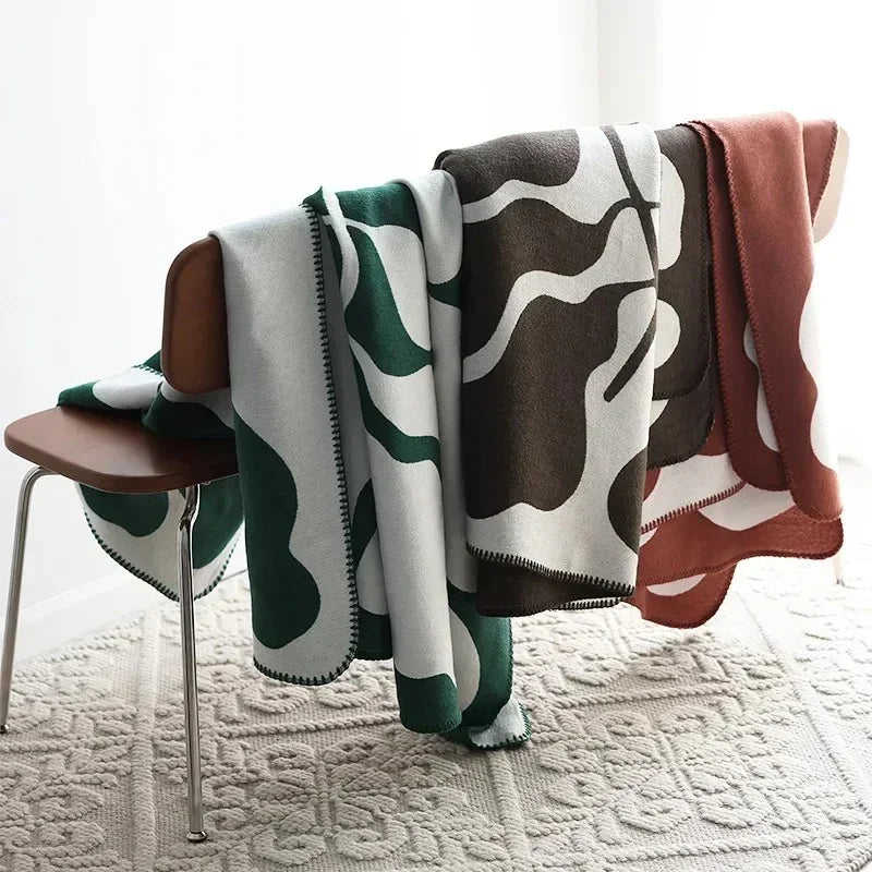 Three knit throw blankets in green, brown and rust, each with the same off-white abstract pattern, hung together over a wooden bench.