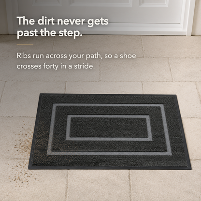 Black door mat on paving with grit caught down between the ribs and a scatter of grit on the clean paving beside it.