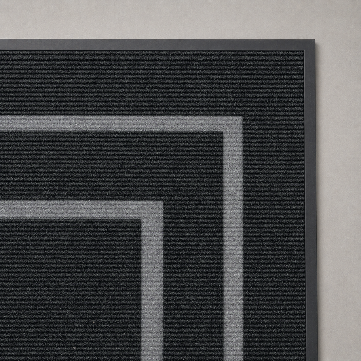 Close-up of the corner of the black door mat on a plain gray background, with both pale gray bands turning the corner, the ribbed pile running parallel to the long edge and the rubber surround along the outer edge.