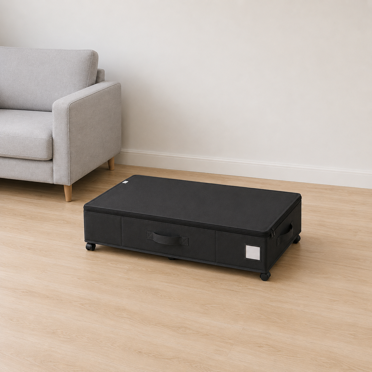 Black under bed storage box on castors, lid zipped shut, on pale oak floorboards beside a light gray sofa in a living room.