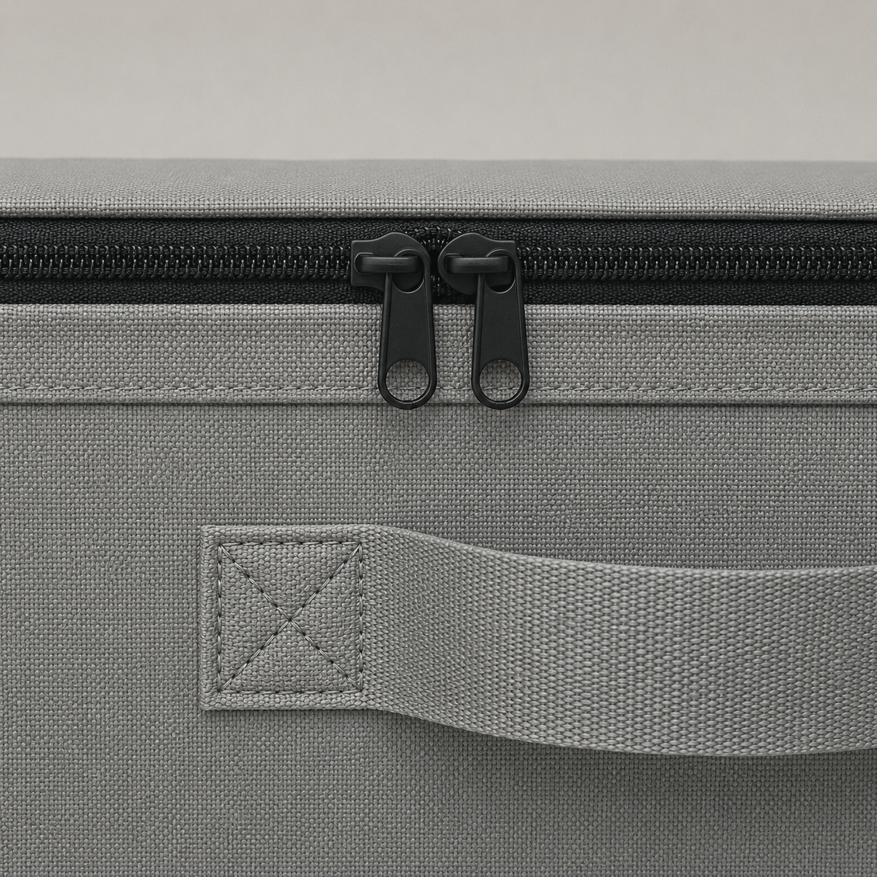 Close-up of the zipper and its twin pulls along the top rail, with a webbing side handle stitched to the gray fabric below.