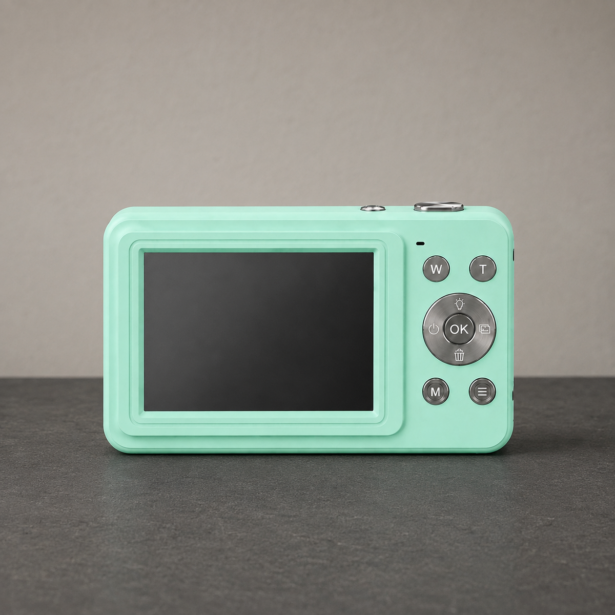Green compact digital camera standing on a dark slate hallway counter against a plain grey wall, seen square on from the back, the screen switched off and blank, showing the W and T zoom buttons, the silver ring around the OK button, and the M and menu buttons