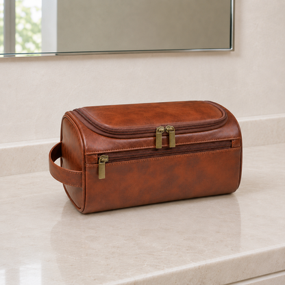 Brown hanging toiletry bag closed on a bathroom counter below a mirror, with warm gold-toned zip pulls and an end handle