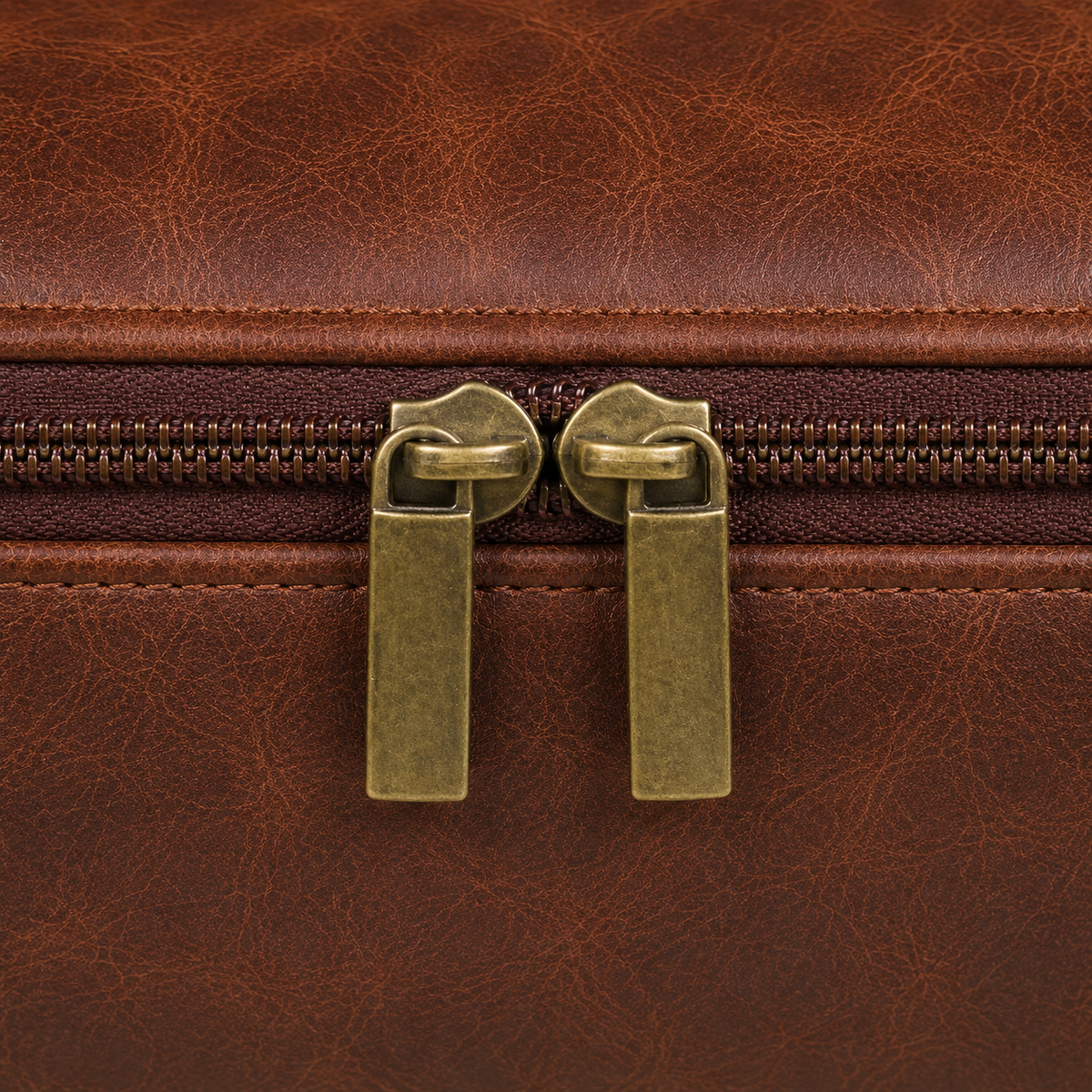 Close view of the two warm gold-toned zip sliders and flat pull tabs meeting on the top zip of the brown bag