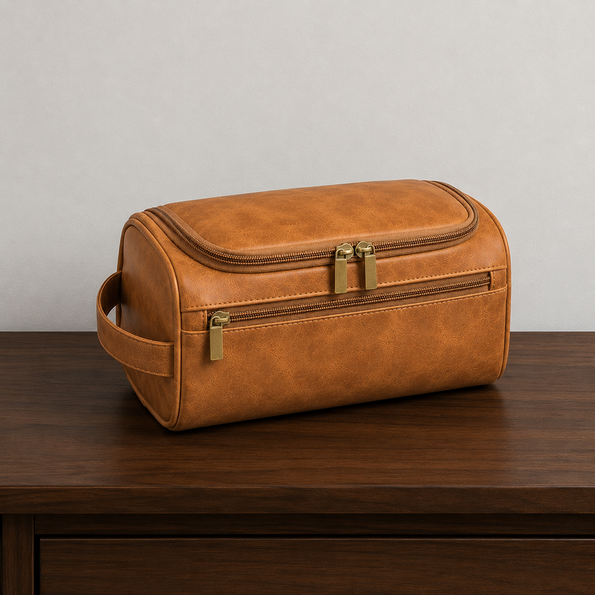 Light Brown hanging toiletry bag closed on a dark walnut hallway console against a plain grey wall, seen from the front left, with warm gold-toned zip pulls and the strap handle on its left end