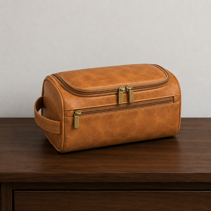 Light Brown hanging toiletry bag closed on a dark walnut hallway console against a plain grey wall, seen from the front left, with warm gold-toned zip pulls and the strap handle on its left end