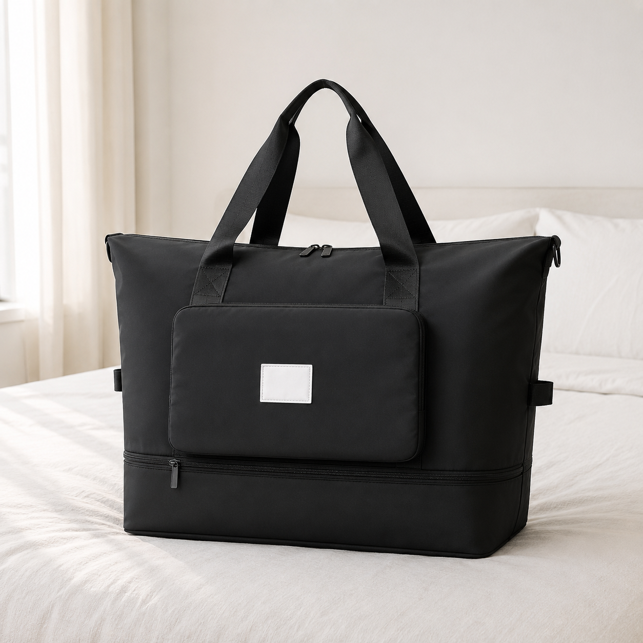 Black foldable travel bag on a hotel bed in daylight, with twin webbing carry handles and a front pocket panel