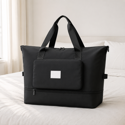 Black foldable travel bag on a hotel bed in daylight, with twin webbing carry handles and a front pocket panel
