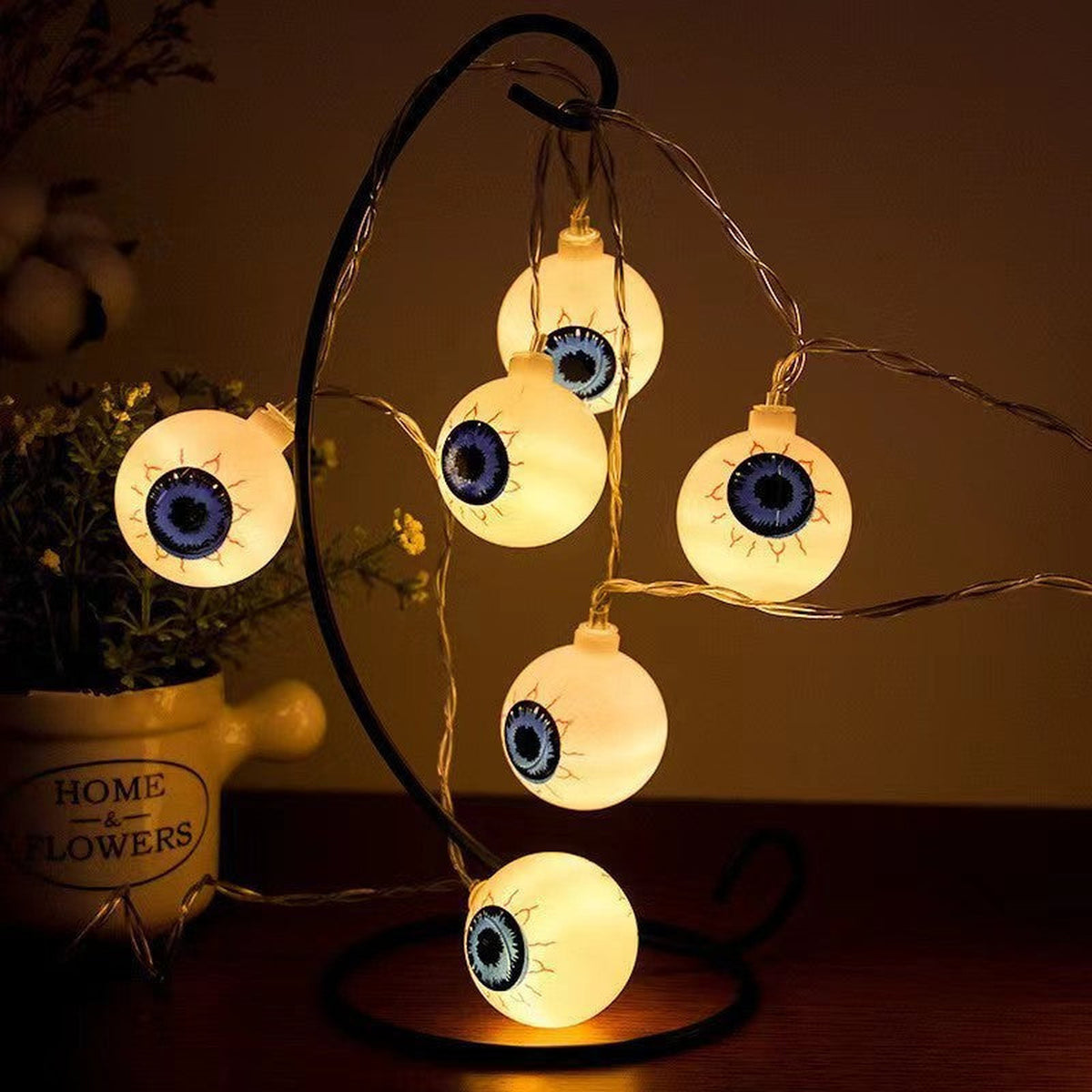 Eyeball string lights lit on a black iron hook stand beside a jug of flowers