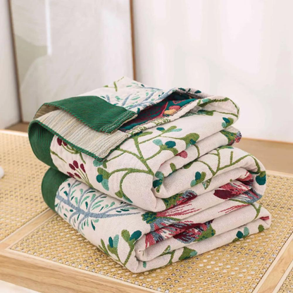 Cream wildflower blanket folded in a stack on a cane bench, showing the green band, the oatmeal band and the teal reverse