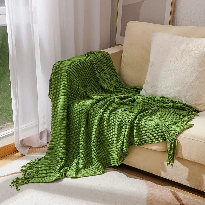 A green bobble knit throw blanket with fringed ends spread over a sofa beside a sunny window.