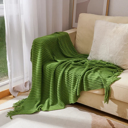A green bobble knit throw blanket with fringed ends spread over a sofa beside a sunny window.